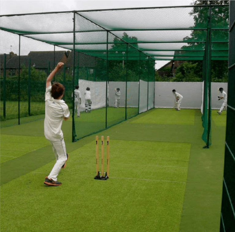 A bowler in white is delivering a cricket ball on a green pitch with wickets set up, in an enclosed netted area.